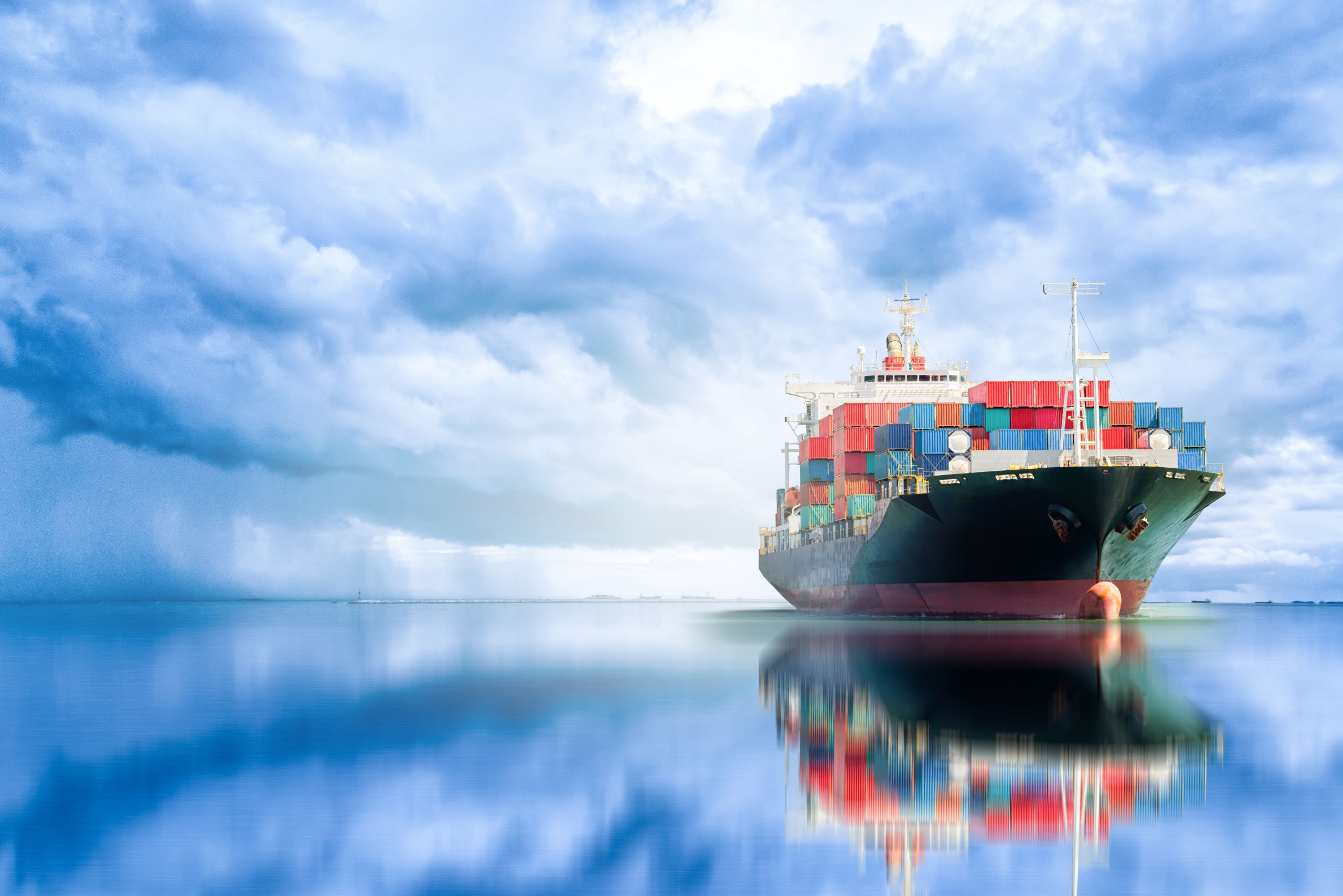 Cargo ship loaded with containers floating on still water under a dramatic cloudy sky.